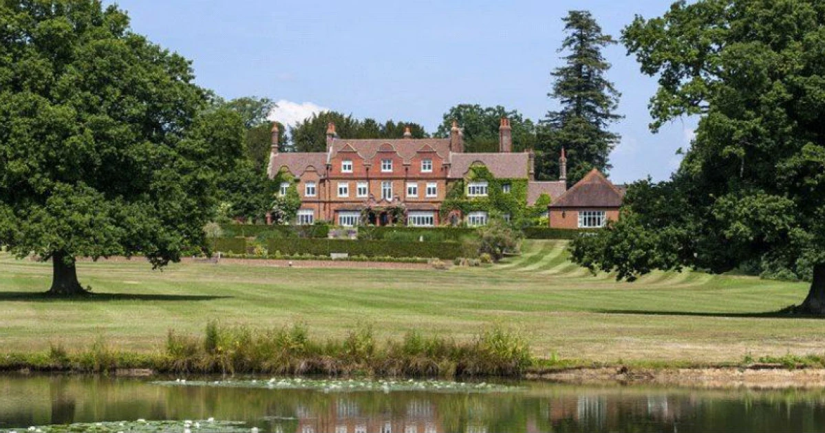 A large English country estate with a red brick mansion, manicured lawns, and a pond in the foreground, framed by tall trees.